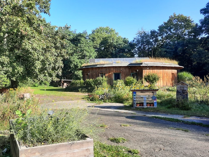 Ein rundes Holzgebäude mit begrüntem Dach steht inmitten eines naturnah gestalteten Gartens mit Hochbeeten, Wildblumen und Bäumen unter blauem Himmel.