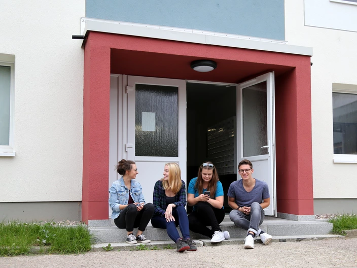 Vier junge Menschen sitzen vor dem Eingang des Studentenwohnheims Ilmenau.