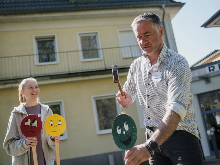 Eine Auszubildende klopft zusammen mit ihrem Auszubildenden Holzschilder mit Smileys in die Erde.