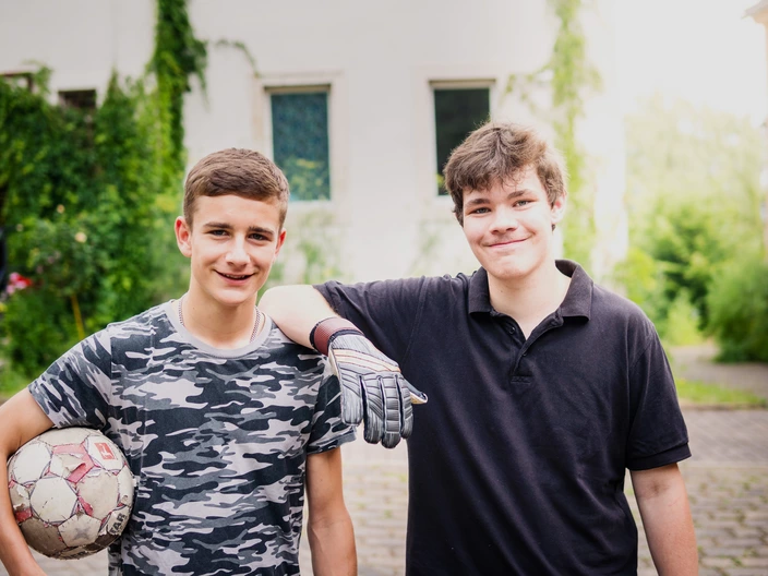 Zwei Jugendliche stehen nebeneinander und lächeln in die Kamera. Ein junger Mann hält einen Fußball unter dem Arm. Der andere junge Mann legt seinen Arm auf der Schulter des anderen ab.