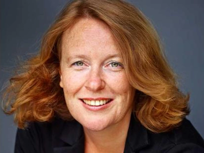 Porträt einer Frau mit schulterlangen, leicht gewellten, rötlich-blonden Haaren. Sie lächelt freundlich in die Kamera und trägt ein schwarzes Oberteil oder Sakko. Der Hintergrund ist einfarbig dunkel, wodurch das Gesicht deutlich hervorgehoben wird. Die Aufnahme wirkt professionell und sympathisch