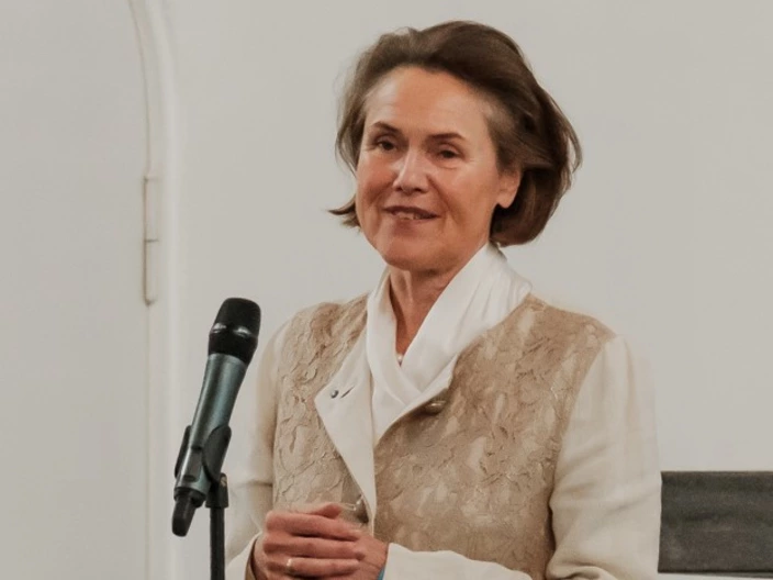 Eine Frau mittleren Alters mit kurzen braunen Haaren spricht in ein Mikrofon. Sie trägt eine helle Bluse mit weißem Kragen und eine beigefarbene, gemusterte Weste. Sie steht vor einer weißen Wand und wirkt freundlich und ruhig. Das Bild scheint bei einer Veranstaltung oder Rede aufgenommen worden zu sein.