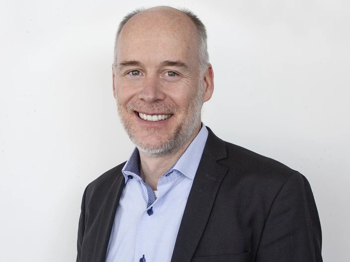 „Porträt eines freundlich lächelnden Mannes mit lichtem Haar und grauem Bart. Er trägt ein schwarzes Sakko über einem hellblauen Hemd mit dunklen Knöpfen. Der Hintergrund ist schlicht und weiß, wodurch der Fokus klar auf der Person liegt. Die Aufnahme wirkt professionell und sympathisch.“