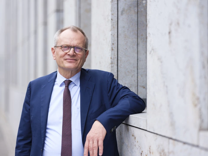 Porträt eines älteren Mannes mit Brille im blauen Anzug, hellblauem Hemd und weinroter Krawatte. Er steht lächelnd an einer hellen Natursteinfassade im Freien und stützt sich mit einem Arm locker auf die Mauer. Die Aufnahme wirkt freundlich, professionell und urban. Im Hintergrund sind die klaren Linien der modernen Architektur unscharf zu erkennen.