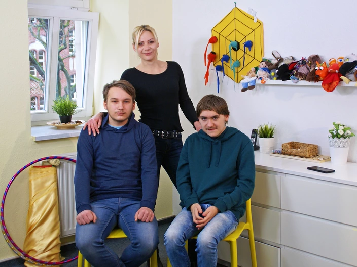 In einem Zimmer mit ein paar Spielsachen steht Kati hinter ihren beiden Söhnen, die vor ihr auf Stühlen sitzen. Spenden helfen Familien in herausfordernden Zeiten die richtige Unterstützung zu bekommen.