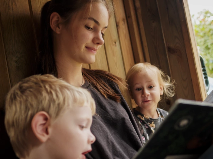 Eine Erzieherin liest zwei kleinen Kindern in einem Baumhaus sitzend etwas vor. Spenden helfen Kindern in Kitas Geborgenheit zu schenken.