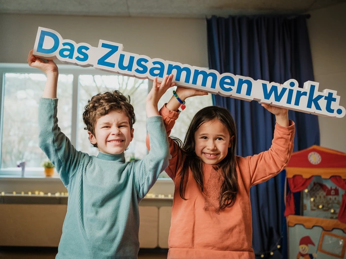Ein Junge und ein Mädchen halten grinsend das CJD Motto "Das Zusammen wirkt." mit ihren Händen über ihren Köpfen. Eine Testamentsspende stärkt frühkindliche Entwicklung und Bildung in den Kindergärten im CJD.