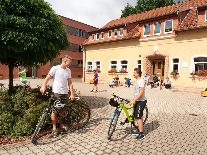 In einem Hof stehen zwei Jugendliche mit Fahrrädern neben einem Baum. Im Hintergrund sind zwei Gebäude zu sehen vor denen mehrere Menschen sitzen und stehen. Spenden stärken die Persönlichkeitsentwicklung von jungen Menschen in Kinder- und Jugendhilfeeinrichtungen des CJD.